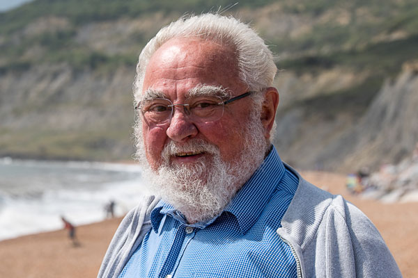 Denys Brunsden, a white gentleman with white hair and beard in a blue shirt, is standing in front of the Jurassic Coast cliffs smiling at the camera. 
