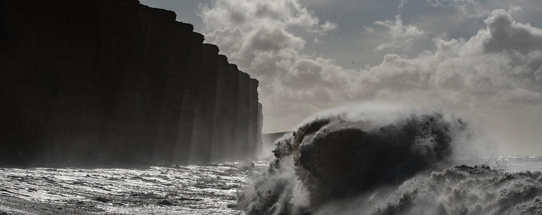 A large storm wave breaks in the sea in front of the cliffs of West Bay. The image is grey and stormy.