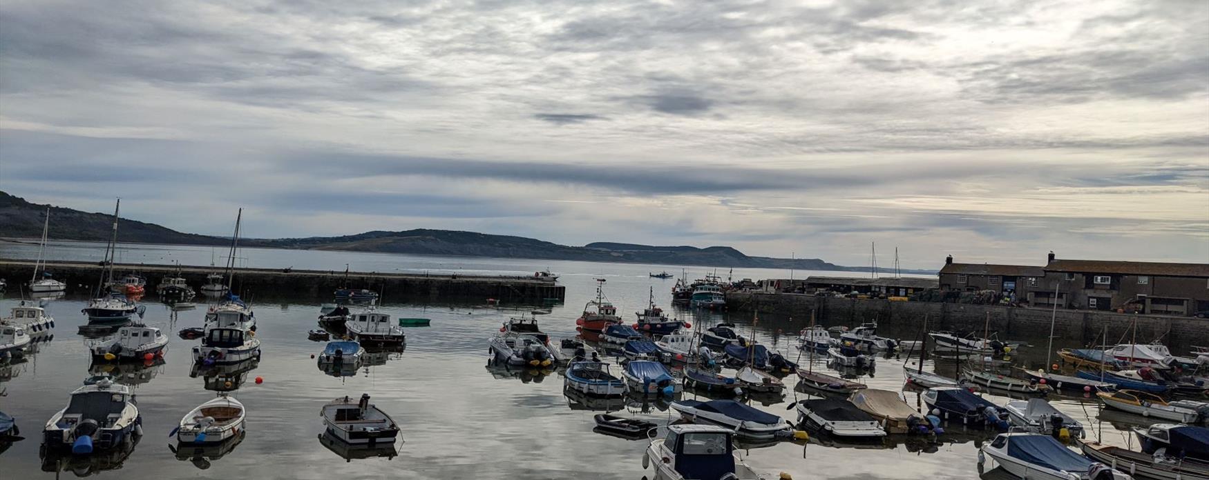 Small boats in a marina at Lyme Regis harbour. The sky is cloudy creating a dramatic sky which is reflected on the sea and the sun is glistening off the sea underneath the boats. The Cliffs of the Jurassic coast extend off away from the image into the distance.