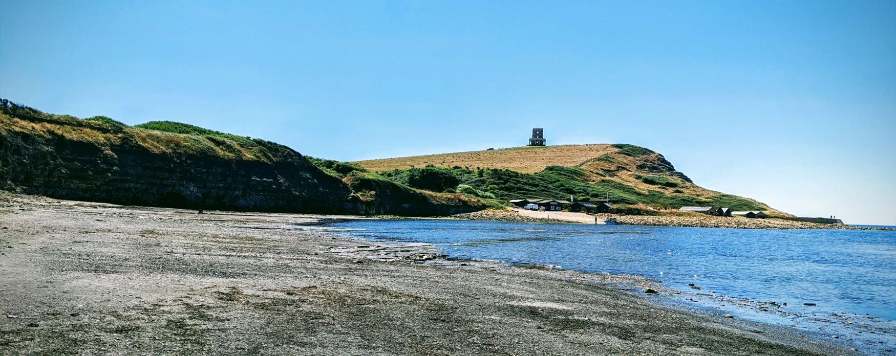 A view of Clavell Tower on top of a grassy cliff at Kimmeridge. The beach the photographer is on has pebbles and large mudflat areas. The sea to the right of the image is bright blue and the weather is calm.