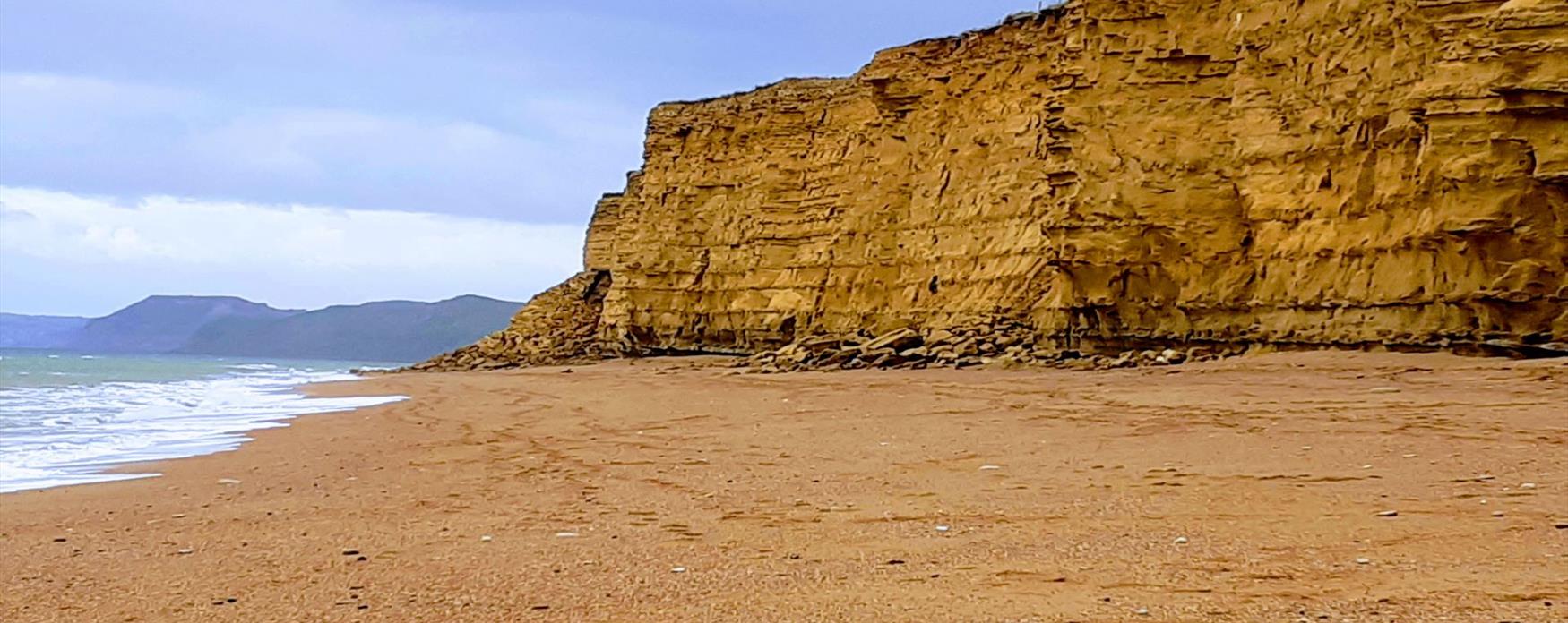 A view of the sandy golden cliffs of Burton Bradstock, slightly contrasting against the browner small pebble beach. The sea to the left of the image is bright blue but with clouds overhead.