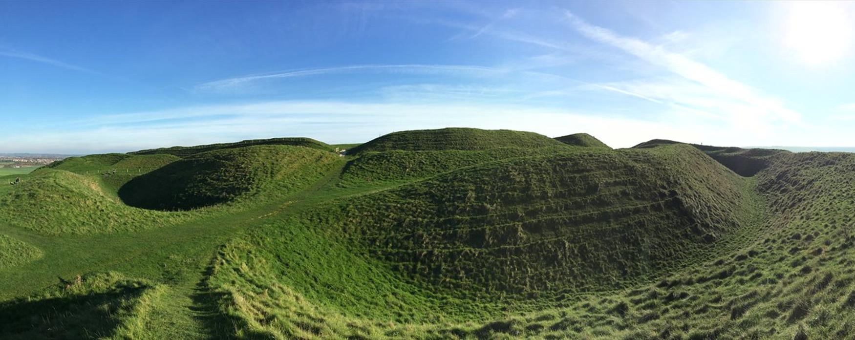 Maiden Castle Hillfort. Large, green, grassy raised earthworks set against a blue sky.