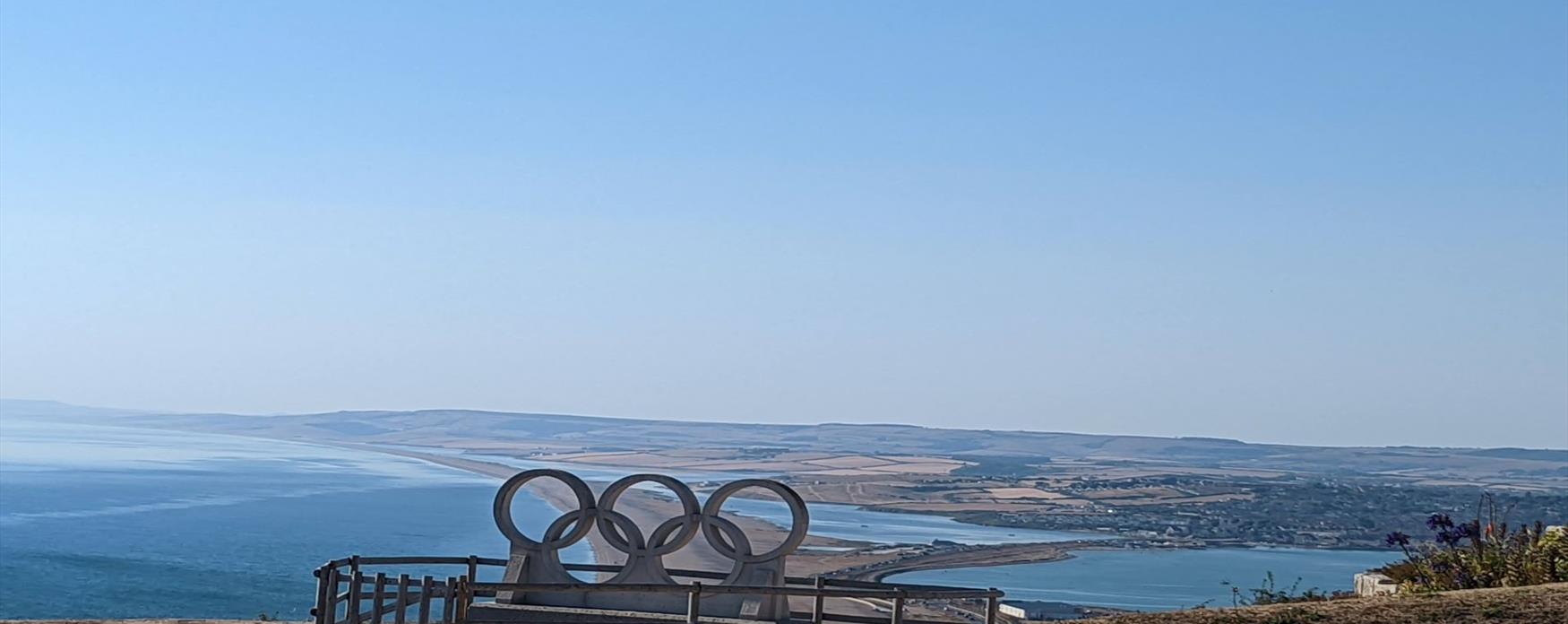 A view of the a long thin strip of beach with water either side extending away from the photographer. A stone statue of the Olympic rings is in the foreground. The built up area of Portland and Chesil is visible to the right of the image.