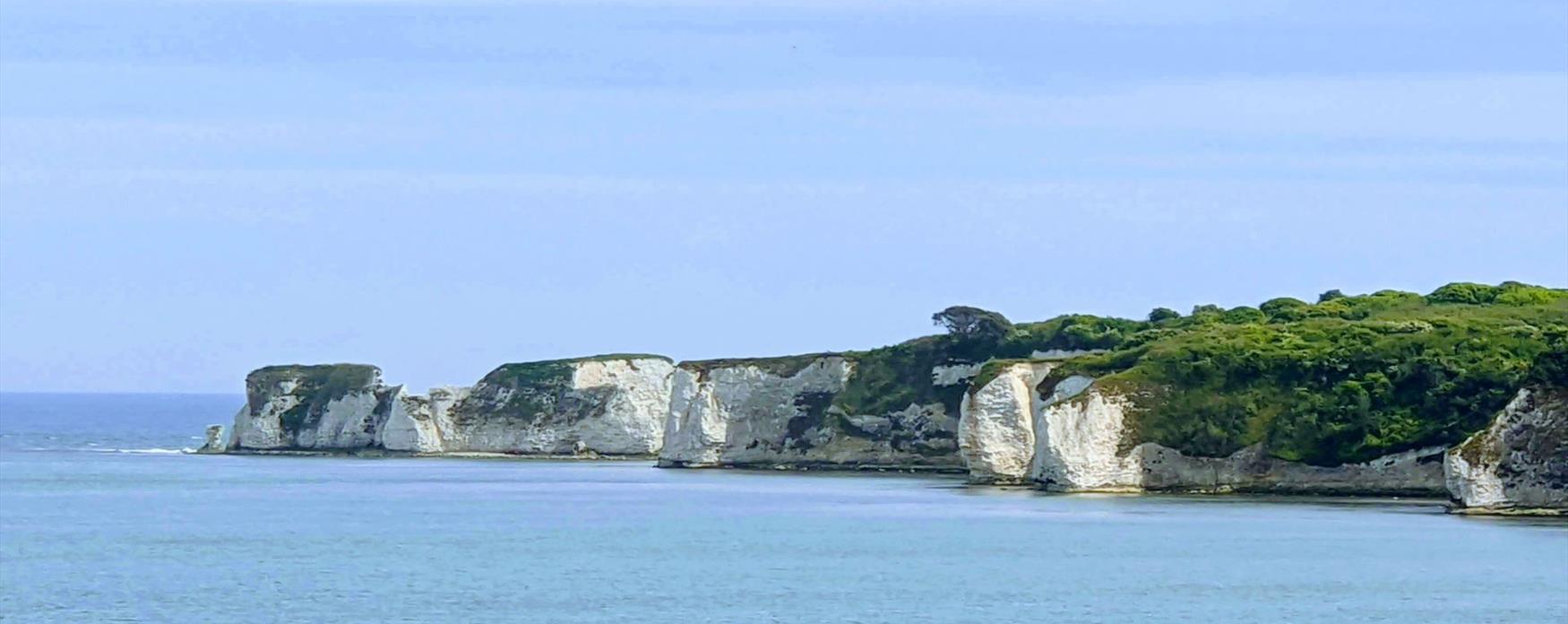 A distant view across the bright blue sea of large white chalk cliffs, topped with green grass, ending in the sea stacks of Old Harry Rocks. The sea is bright blue and glistening in the sun.