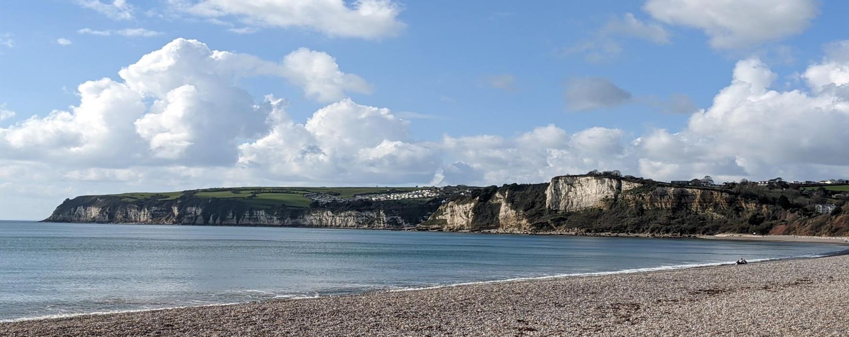 A sweeping view of the landscape from Seaton to Salcombe Hill. The cliffs are predominantly of the white chalk cliffs of Beer, with red cliffs in the foreground. The beach where the photographer is stood is a pebble beach. The sun is shining off the bright blue sea on the left side of the image.