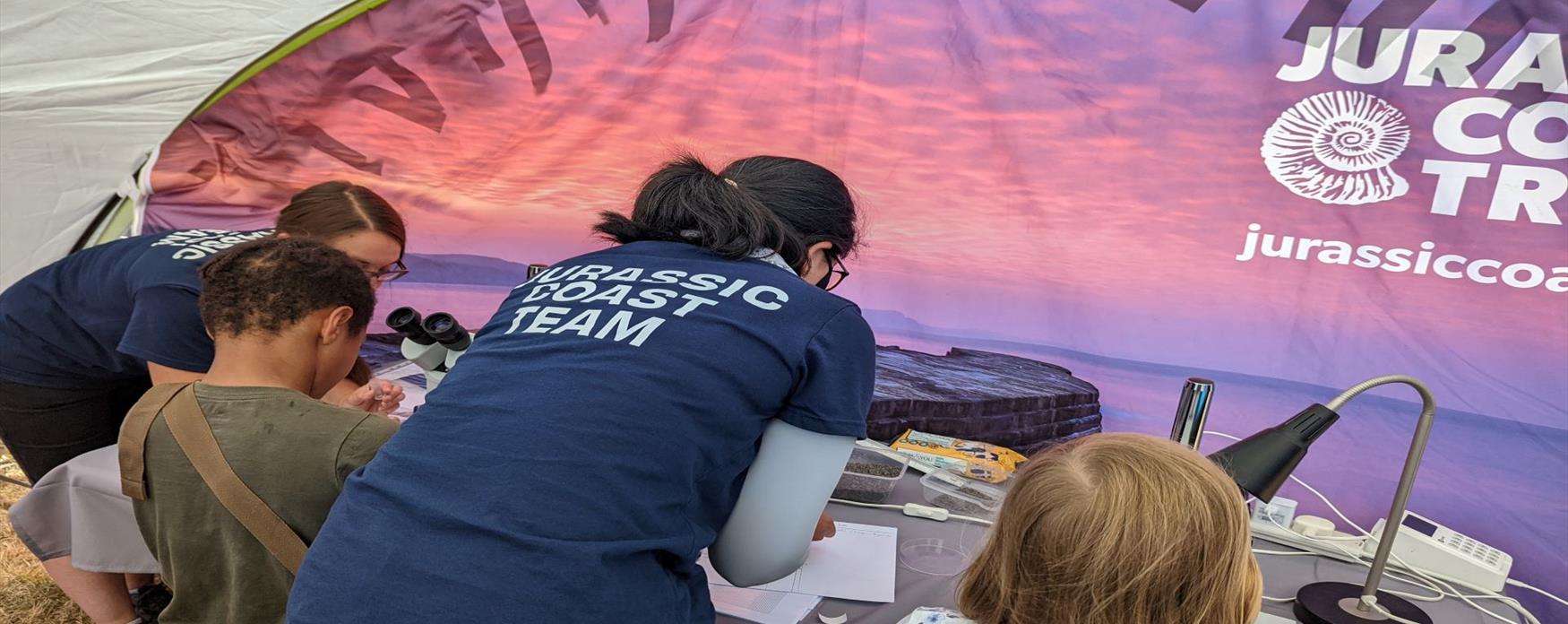 Two children are seated at a table in front in a Jurassic Coast Trust gazebo, being shown by two people in blue Jurassic Coast Trust t-shirts, how to find micro-fossils on petri dishes under microscopes. The gazebo panel in front of the table is purple and pink with a Jurassic Coast Trust logo in the top, right corner.