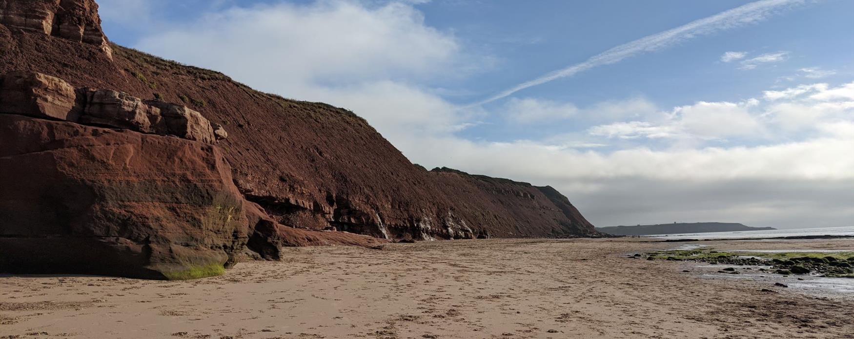 A light sandy beach scene with bright red cliffs on the left. The cliffs have a vibrant red colour with green grass capping them and succulent plants growing intermittently along the cliff. The sea is bright blue and appears slightly on the right side of the image. There are some light white fluffy clouds in the sky over the cliffs.